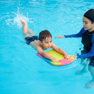 A child holds a kickboard and kicks through the water with an instructor guiding.