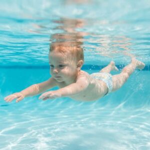 A young child swims confidently and independently just beneath the sunlit surface of a clear blue pool.