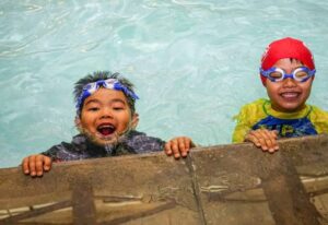 Two young children smile and hold onto the edge of a swimming pool during a group class.