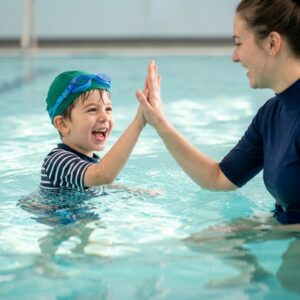 A young boy beams with confidence as he high-fives his swim instructor in the pool.