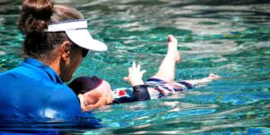 A toddler practices floating on their back with the gentle assistance of a swimming instructor.