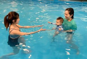 A toddler walks toward an instructor in a shallow indoor pool during a structured swim lesson.