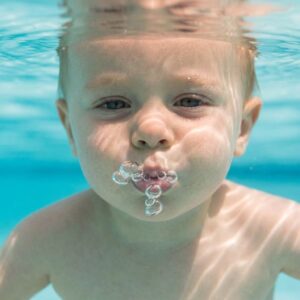 A close-up view of a child submerged underwater in a pool, actively blowing a stream of bubbles.
