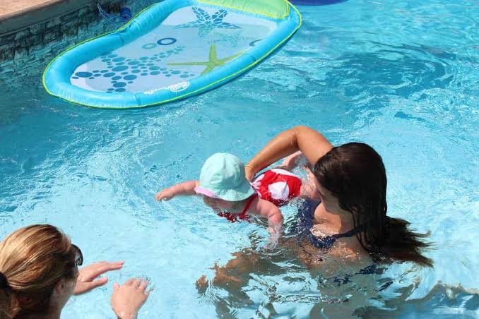 A smiling toddler receives gentle support from a swimming instructor during a water safety lesson.