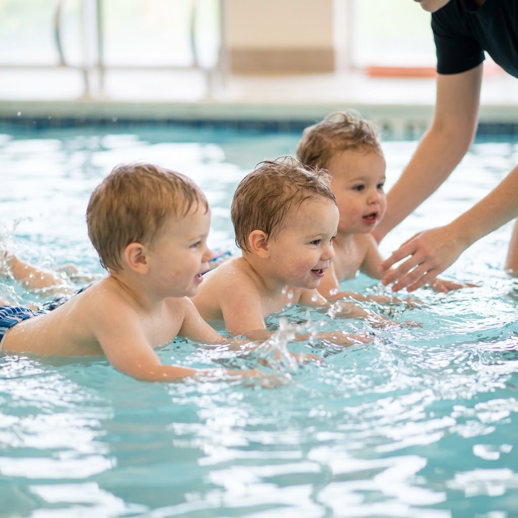 Three joyful toddlers paddle and kick in clean indoor pool water during a small group swim lesson with instructor hands nearby.